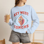 Key West Conchs Crewneck Sweatshirt in light blue with the grinning striped conch shell mascot inside a teal diamond outline on the front, worn by model