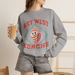 Key West Conchs Crewneck Sweatshirt in sport grey with the grinning striped conch shell mascot inside a teal diamond outline on the front, worn by model
