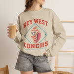 Key West Conchs Crewneck Sweatshirt in sand with the grinning striped conch shell mascot inside a teal diamond outline on the front, worn by model