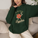 Macon Peaches Hoodie in forest green with the cartoon peach mascot tossing a baseball on the front, worn by model